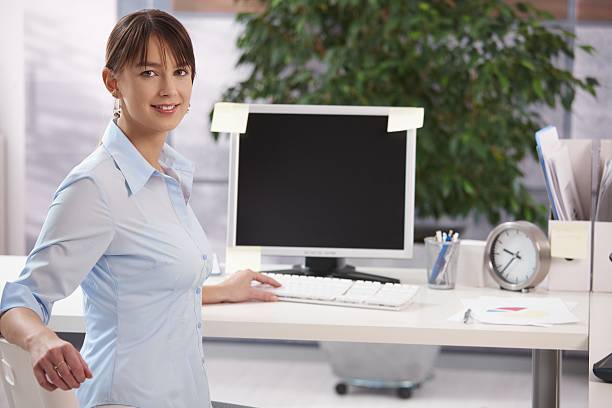 Young female office worker sitting at desk in office, working on computer, looking back and smiling to camera. Blank space on monitor.