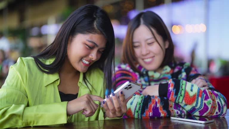 Two multi-ethnic female friends looking at mobile smart phone and laughing in café