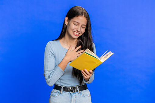 Happy relaxed smiling Caucasian young woman reading funny interesting fairytale story book, leisure hobby, knowledge wisdom, education, learning, study, wow. Teenager girl isolated on blue background