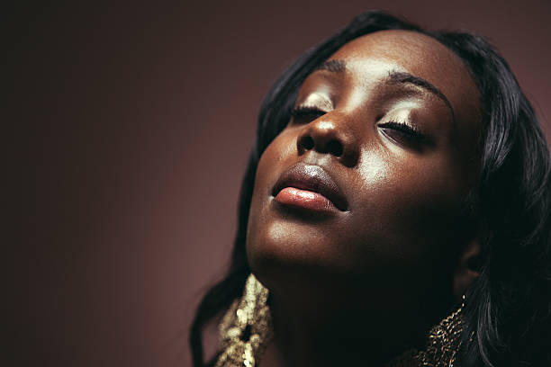 Beautiful Young African Woman An attractive young adult African woman looks upward with eyes closed, a look of peace and contentment on her face as she dreams / prays.  Horizontal studio shot on a brown background. drop earring stock pictures, royalty-free photos & images