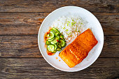 Seared salmon steak with white rice and sliced cucumber on wooden table
