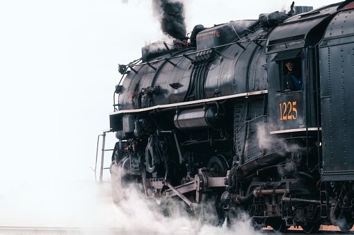 Owosso, United States – December 06, 2021: A black and white steam locomotive engine chugging along a winding track on a cloudy day