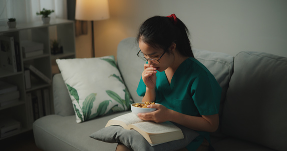 Portrait of Young Asian woman healthcare worker in medical scrubs wearing eyeglass enjoy reading a book and eating cookie on comfortable sofa at home,Wellness lifestyle in scrubs