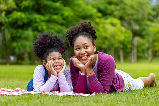 African American mother and young daughter are lying down after having a summer picnic in public park for weekend leisure and happiness