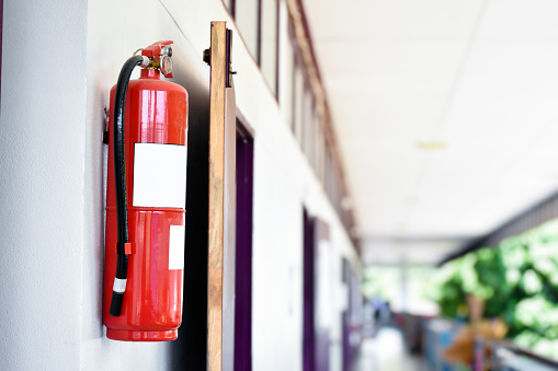 A red fire extinguisher is installed on a white cement wall in the front porch of the building to be used to extinguish a fire in the event of a building fire.