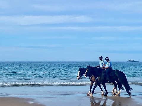 Horizontal seascape of two Australian police officers and horses patrolling a popular summer beach at Byron Bay NSW Australia