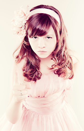 Portrait of a young girl dressed in a pink dress pointing her finger up towards the camera with an angry look on her face.