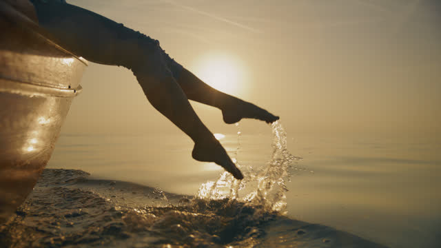 SLO MO Graceful Splashes: A Woman's Feet Splash the Water While Sitting on the Bow of a Sailing Boat at Sunset