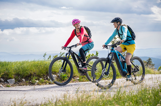 Two happy mid adult women wearing protective helmets riding electric mountain bikes together. Healthy lifestyle concept.