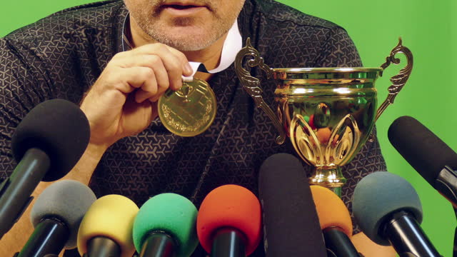 champion speaking with a medal and cup at a press conference in front of microphones - green background