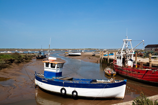 Fishing boats beside the small quay at Brancaster Staithe, Norfolk. Main focus on blue and white fishing boat tied to posts on land. A more open red fishing boat is on its left. There is a smaller row boat next to the red boat. Clear skies.