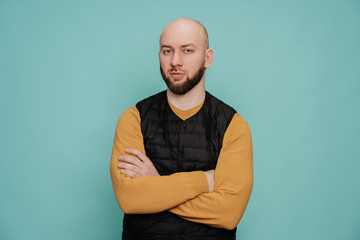 Bald caucasian bearded man in casual clothes standing hands folded on chest looks at camera with focused calm face against turquoise studio background with copy space.