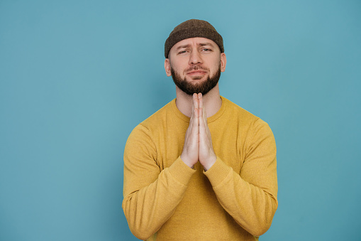 Studio shot of handsome unshaven male wearing yellow sweater with confusing expression, asking for luck, looks puzzled, isolated over blue background. Beseeching bearded man indoor.