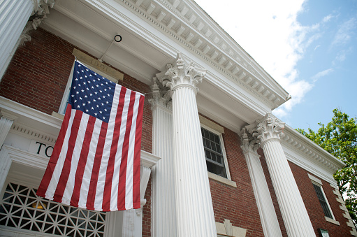 American flag hangs between the white columns of a traditional colonial style town hall building