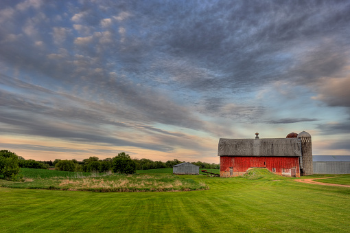 A country Red barn and summer evening cloudscape in rural Minnesota. This photo includes farm silos and sheds with green grass in the foreground. I used a wide angle 14-24 mm lens to help with the framing of this landscape shot.