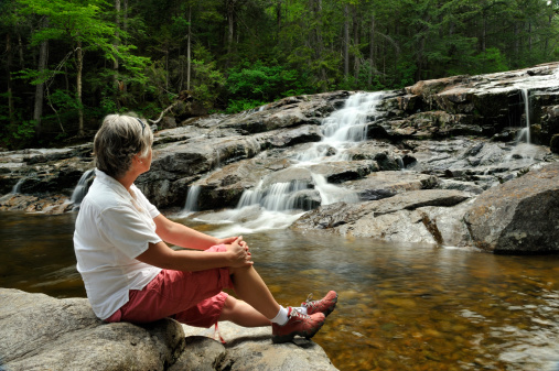 This is a horizontal, color, royalty free stock photograph of a young, caucasian, 10 year old American boy on summer vacation in the Catskill Mountains in upstate New York, United States. He jumps from rock to rock to cross the wooded stream in the forest. His uncle is in the foreground putting a log in place to make the crossing easier for the boy. Green trees fill the background. Photographed with a Nikon D800 DSLR camera.