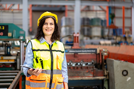 portrait of smilling engineer worker woman in safety vest and helmet standing with digital tablet, people working at industrial manufacturing factory inspect repairing equipment of production machine
