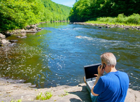 A man telecommuting by a beautiful mountain river calling into the office.See more of my laptop images at