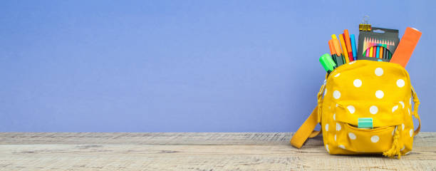 istock Yellow backpack with white polka dots with different colorful stationery on table. Purple background. Back to school. Banner design. Banner design. 1828285939