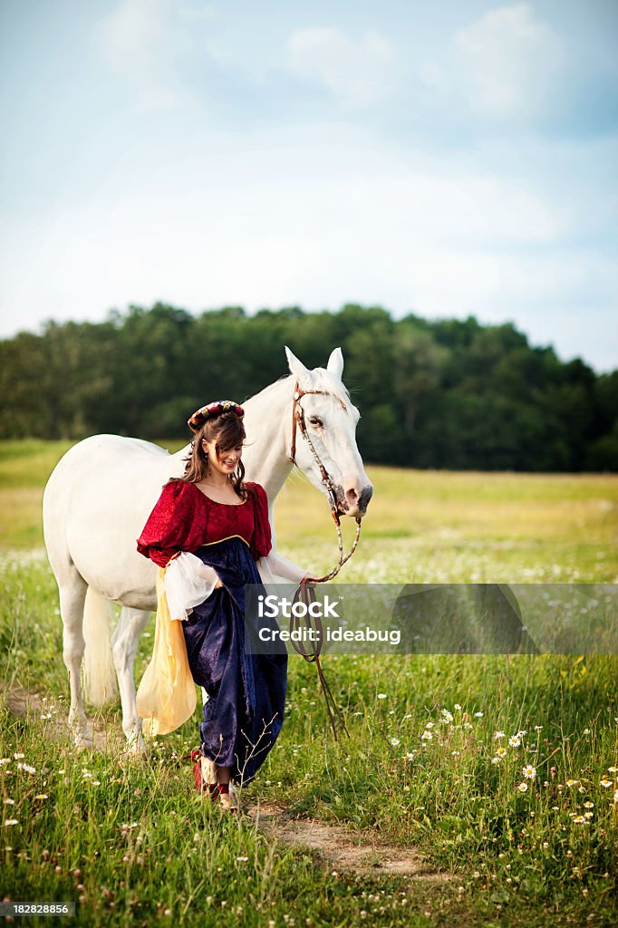 Young Woman Wearing Renaissance Dress with Horse Walking in Field Color photo of a young woman dressed in a renaissance-age, empire dress walking through a field with her white horse on a sunny, summer day. Adult Stock Photo