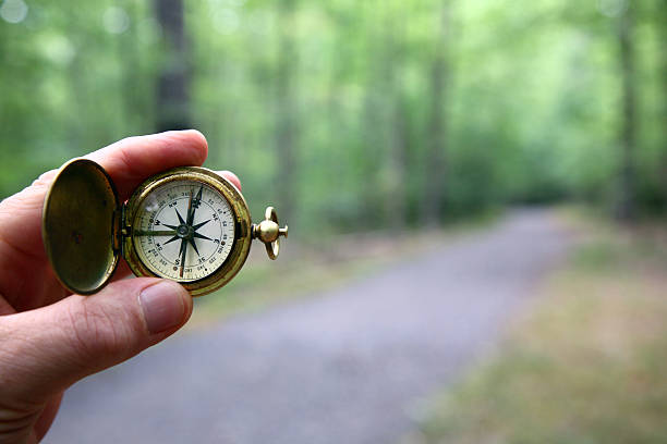 Compass With Blurred Woods Trail Old brass pocket compass held in fingertips with a blurred gravel path and woods in background. north stock pictures, royalty-free photos & images
