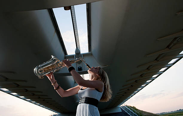 three quarter length side view shot of a young blond woman playing on saxophone standing under bridge at sunset