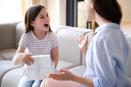 Happy amazed kid opening present box at home. Young mother sitting on sofa and making birthday surprise to her preteen daughter. Woman congratulate child and give positive emotions.