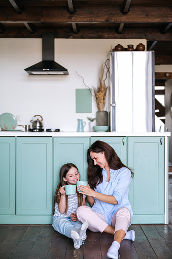 Vertical shot of happy mother holding cup of herbal tea and giving to smell beverage to smiling daughter, sitting on kitchen floor, having breakfast and spending weekend together at home