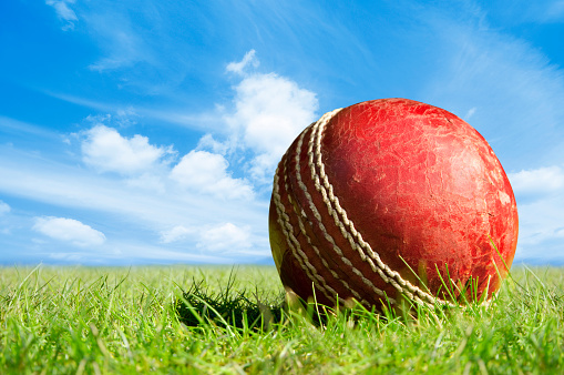 A red leather cricket ball lying on a grass cricket pitch under a blue summer sky. The ball is old and worn and the leather is cracking. It casts a strong shadow into the green grass beside it from the strong sunshine. The blue sky has white clouds in it.