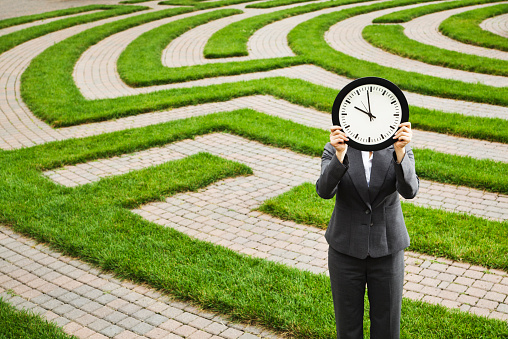 A business woman holding a clock over her face in front of a corporate maze, searching for a solution to her business problem. Concept of business problem solving and management strategy.