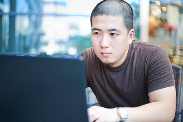 Young man looking intently at his laptop in a cafe An asian young man is using a laptop in a outdoors cafe. crew cut stock pictures, royalty-free photos & images