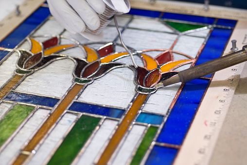 A craftsman wearing protective gloves uses an iron and lead solder to join seams on a copper-foil window project.