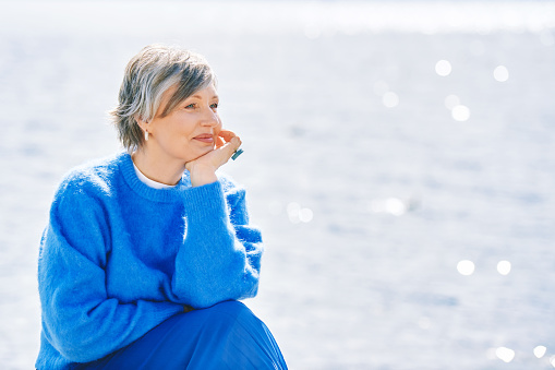 Outdoor portrait of beautiful mature woman relaxing next to lake or sea, wearing warn cozy pullover, healthy lifestyle