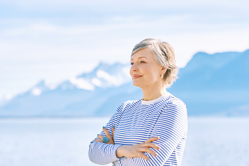 Outdoor portrait of beautiful mature woman posing next to lake, wearing blue stripe t-shirt, healthy lifestyle