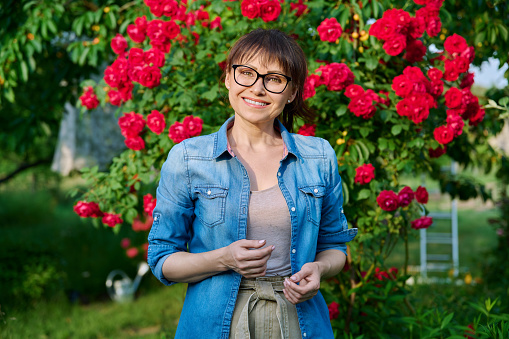 Portrait of middle-aged woman in backyard background blooming red rose bush. Smiling 45 year old female looking at camera. Beauty of nature, people concept