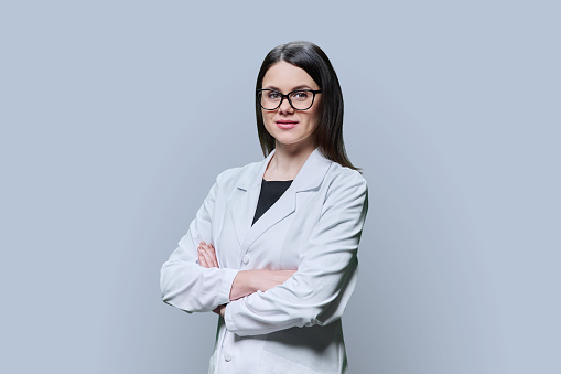Portrait of young confident woman specialist, professional medical scientist, pharmacist in white coat with crossed arms on grey background. Medical staff, occupation, health care, science medicine