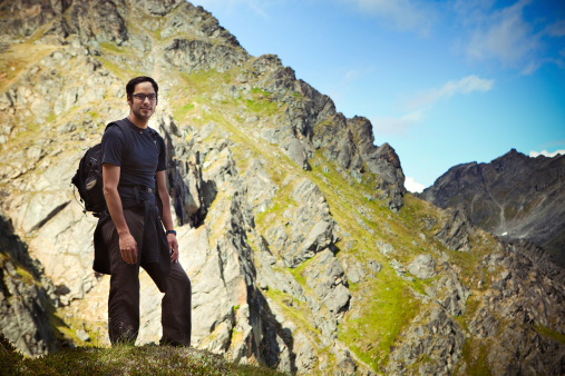 A young native Alaskan man on a hike stops on a grassy ledge to admire the rocky terrain view below.  Horizontal with copy space.
