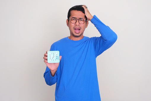 Adult Asian man showing panic expression while holding alarm clock