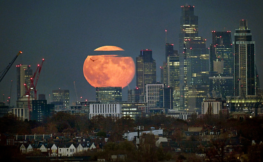 The full moon rises over the distant City of London financial district with foreground suburban homes