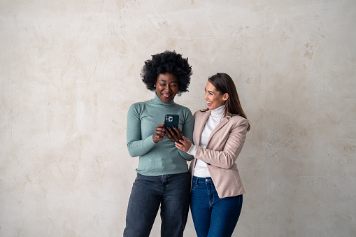 Two cheerful interracial women using mobile phone while standing in a studio.