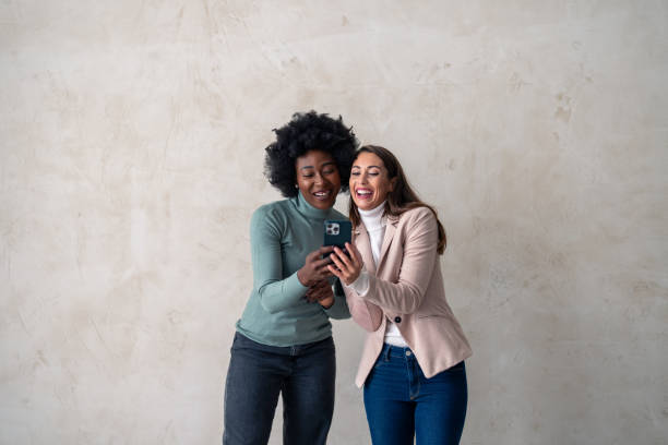 istock Two cheerful interracial women in casual clothing using mobile phone while standing in a studio 1828552075