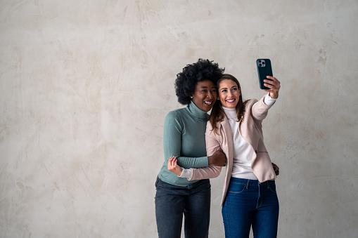 Two happy charming women enjoying themselves while standing together and taking selfies with smart phone.