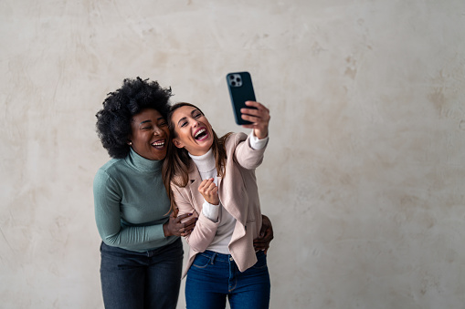 Two multicultural women broadly smiling while taking selfie with mobile phone.