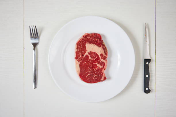 A raw ribeye steak on white plate with knife and fork on a white table