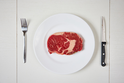 A raw ribeye steak on white plate with knife and fork on a white table