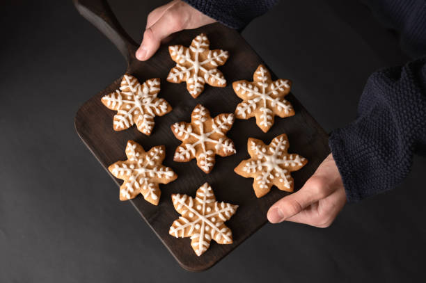 istock Hands holding wooden board with snowflake cookies decorated with sugar icing, on dark background. Traditional Christmas home treat, food. Soft selective focus, lifestyle 1828571939
