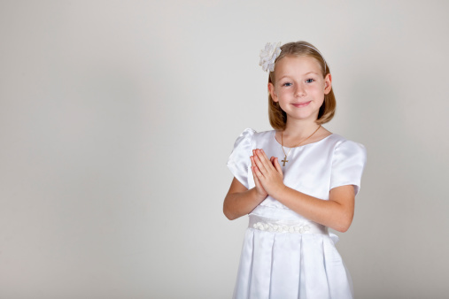 Beautiful little girl in communion dress with praying hands looking at the camera smiling.