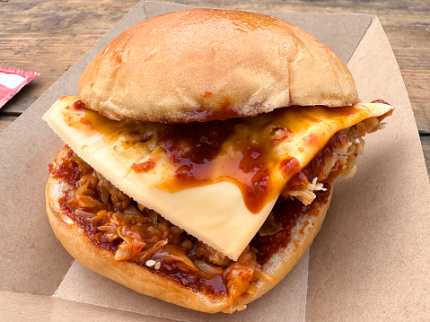 Stock photo showing close-up, elevated view of chicken burger served in glazed, brioche bread bun in cardboard takeaway box. The burger is made with chicken breast, processed cheese slice and Korean sauce.