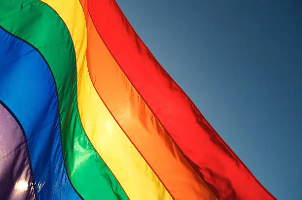 Rainbow flag glows in bright sun waving in blue sky. Some motion blur on top edge of waving flag.