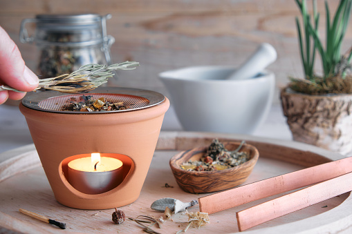 Burning various types of incense like frankincense, myrrh, sandalwood and herbs in an incense burner with tea light - placing a branch of rosemary, jar with incense mixture and mortar in the background. Spring still life, eye level view, wellbeing and spiritual concept